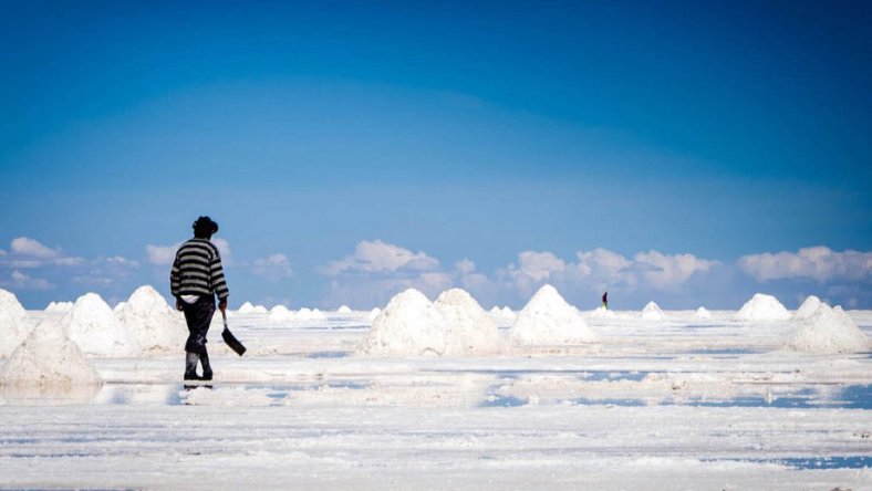 Worker walking across a salt flat with white salt mounds under a blue sky
