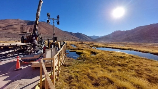 Drilling rig on a wooden platform beside a stream winding through golden highland grassland under bright sun